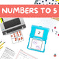 A flat-lay of a children's numbers to 5 learning set, featuring a blue plastic case with number cards, printed worksheets, a printer, and colorful pencils on a white desk. A coral banner at the top reads 'NUMBERS TO 5'.